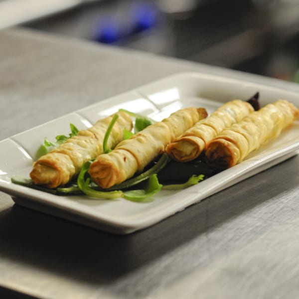 Four golden-brown Mediterranean spring rolls, garnished with fresh greens, on a white rectangular plate.