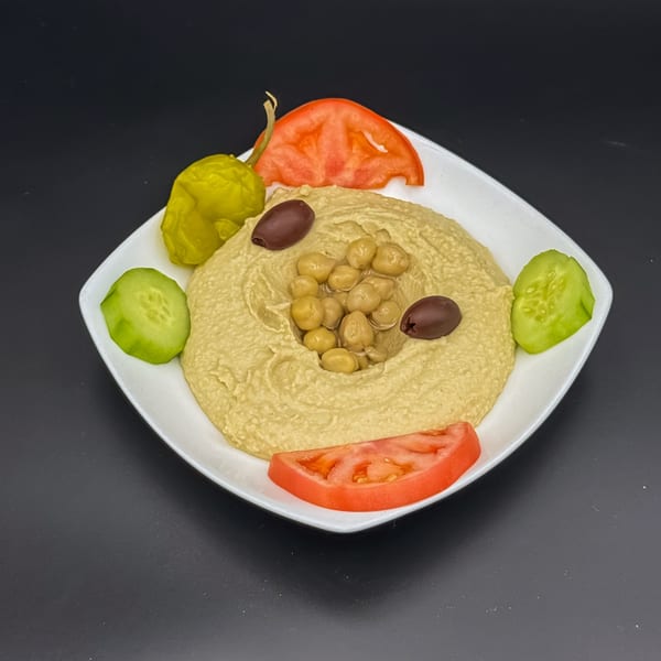 Creamy hummus topped with chickpeas, olives, tomatoes, cucumbers, and a pepperoncini on a white plate.